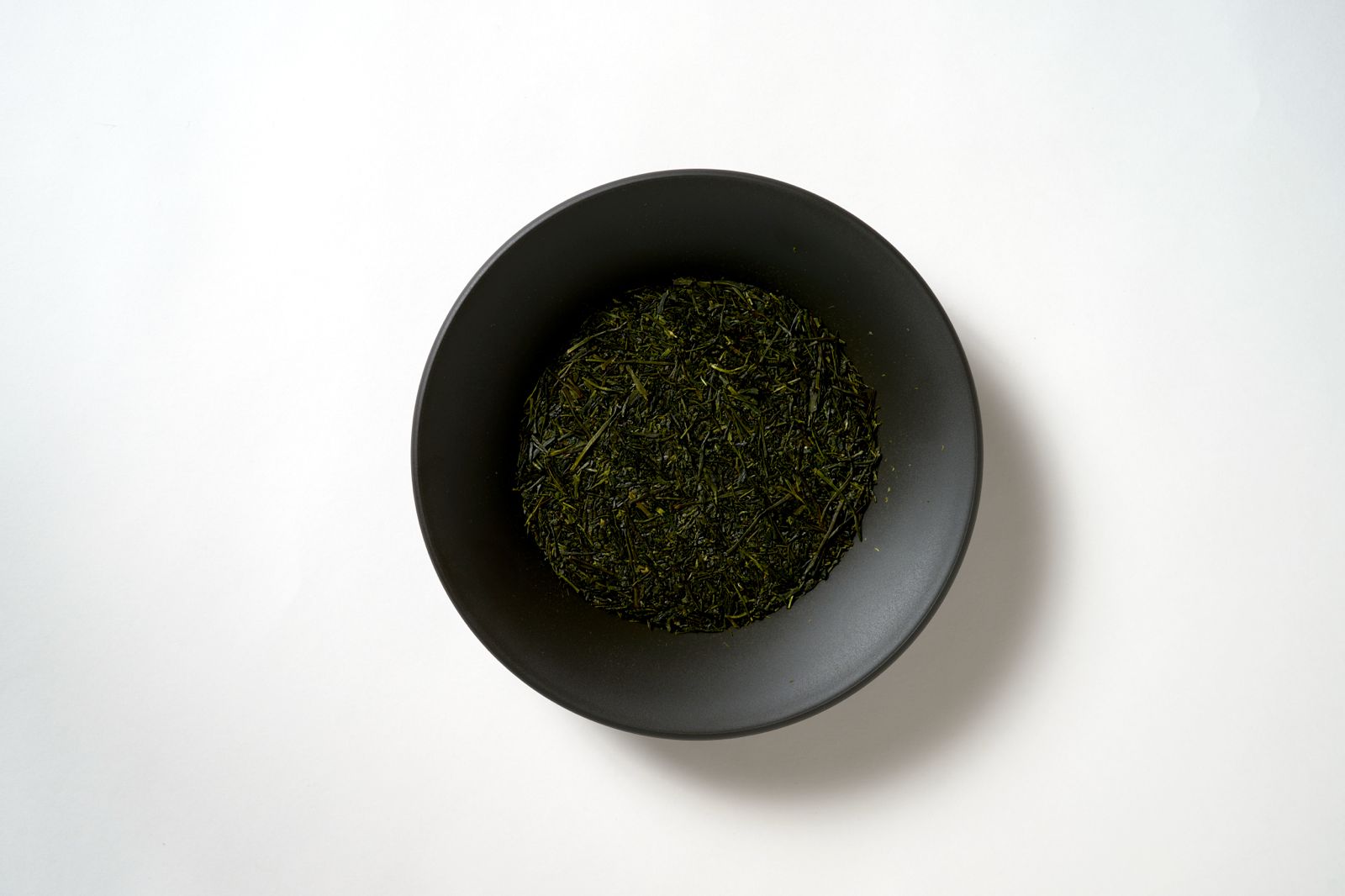 Deep-steamed fukamushi sencha dry leaves in a dark ceramic bowl, showing the fine broken particles characteristic of deep steaming